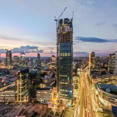 Warszawski wieżowiec w budowie na tle zachodzącego słońca i panoramy miasta.