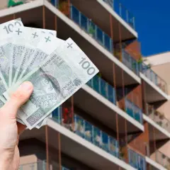 Dłoń trzymająca polskie banknoty 100 zł na tle nowoczesnego budynku mieszkalnego.