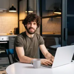 Uśmiechnięty mężczyzna z kręconymi włosami pracuje na laptopie przy stole w kuchni.