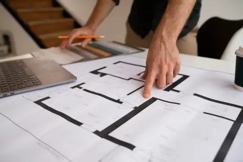 Dłoń wskazująca na plan mieszkania, obok laptop i próbki materiałów.
