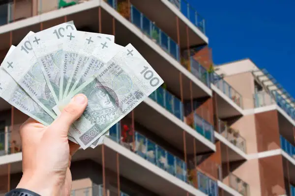 Dłoń trzymająca polskie banknoty 100 zł na tle nowoczesnego budynku mieszkalnego.